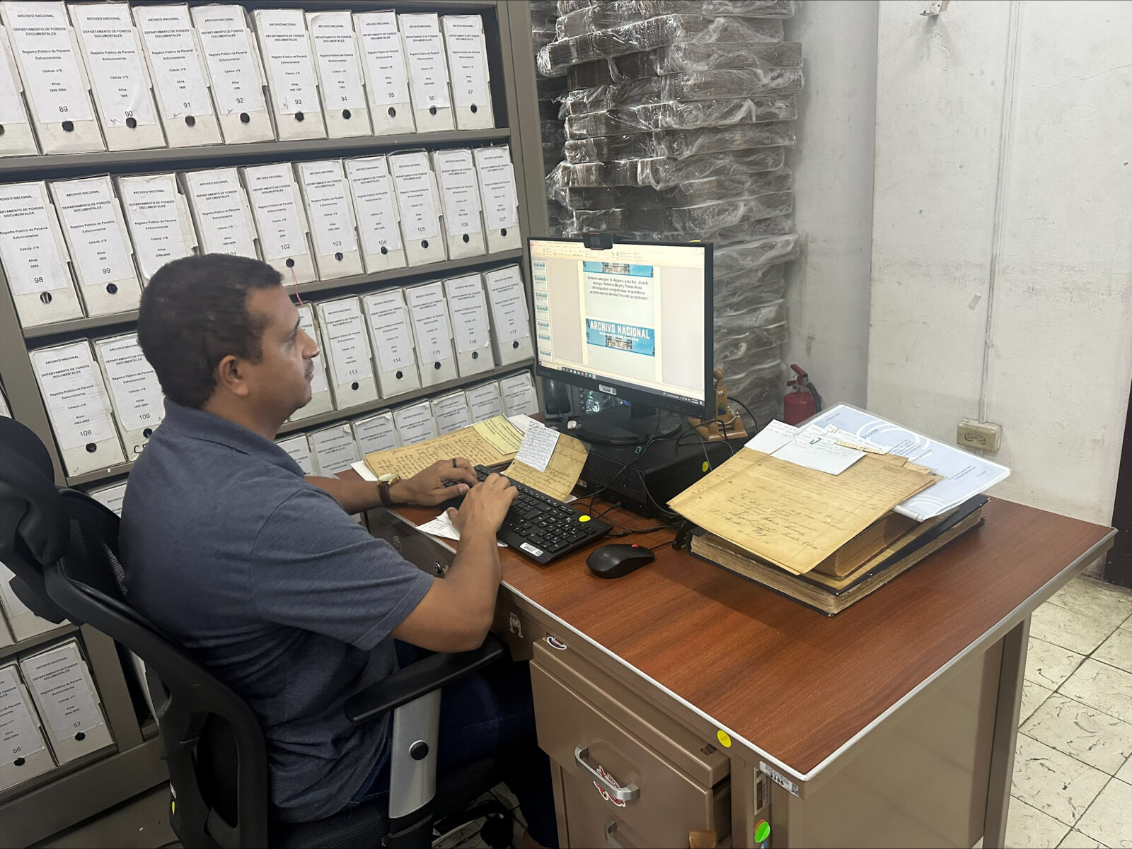 Persona en ordenador rodeado de estanterías con archivadores y documentación